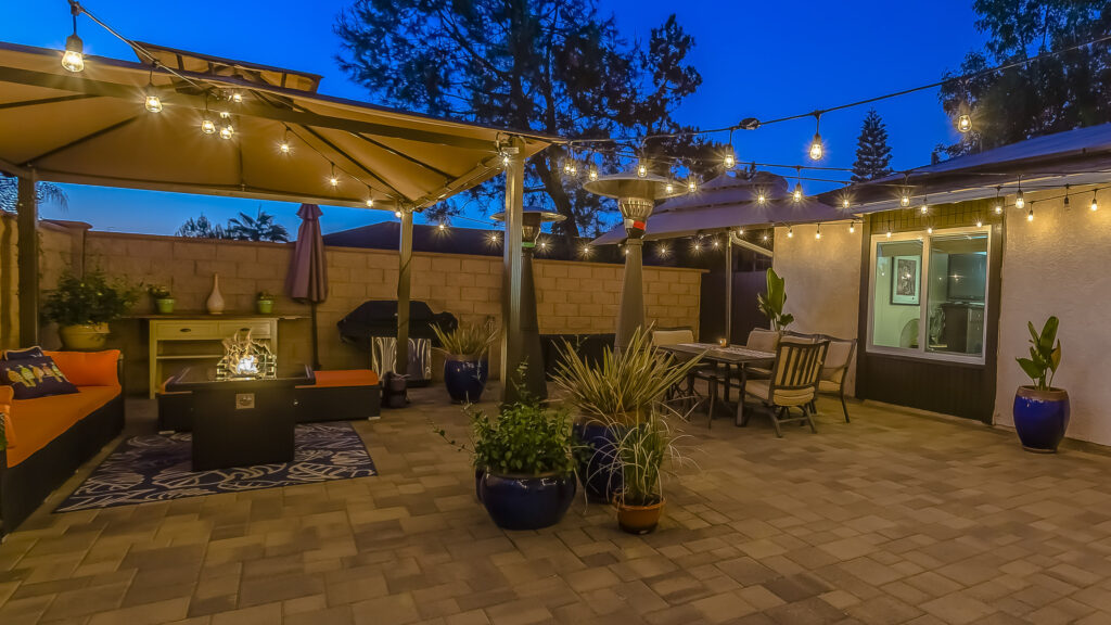 Panorama Patio of home with seating area under a gazebo and dining area under an umbrella. The outdoor space is decorated with potted plants and illuminated by string lights.